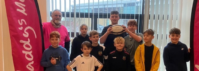 Pupils from Cranbrook Education Campus stood in a conference room at Sandy park, Exeter with two big red flags either side and Exeter Chiefs Coach.